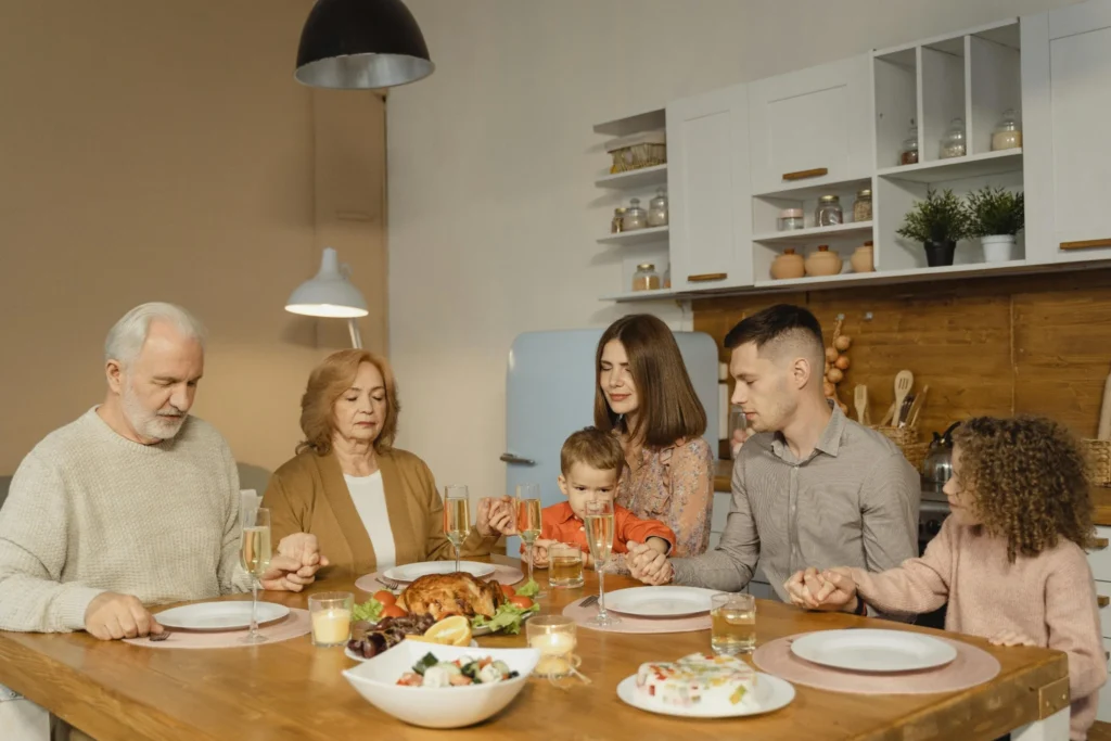 A multi-generational family holds hands and prays together before sharing a meal in a warm, welcoming kitchen.