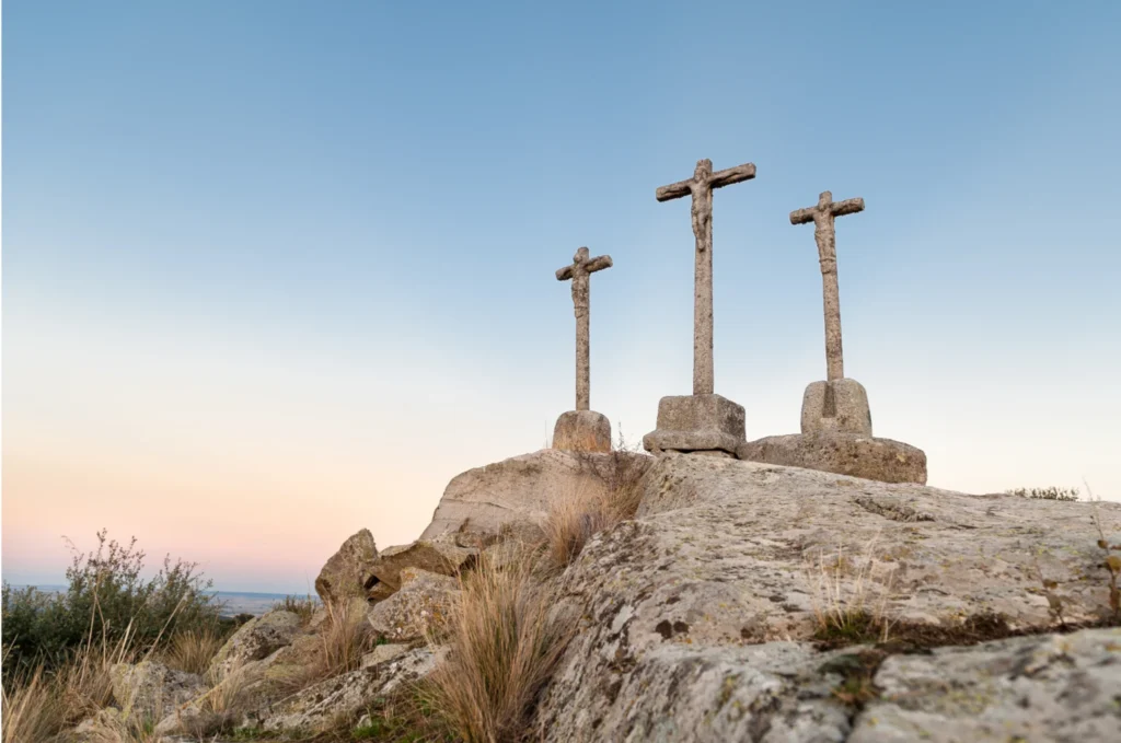 Three stone crosses on a rocky hill against a soft blue sky, representing the crucifixion of Jesus on Good Friday