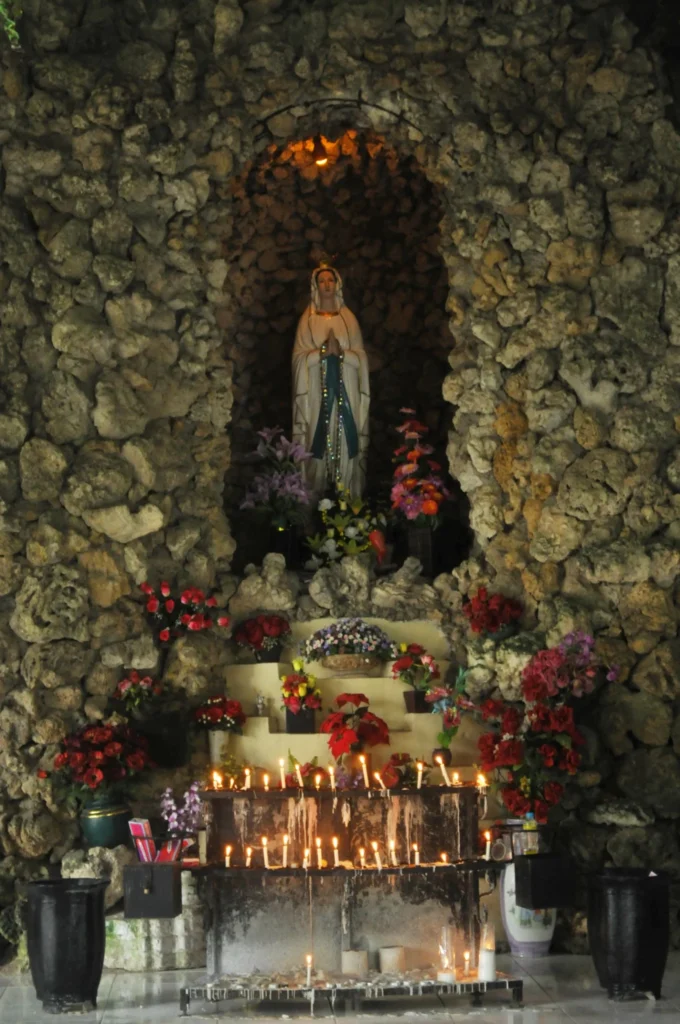 Marian grotto with prayer candles and flowers at a Catholic shrine.